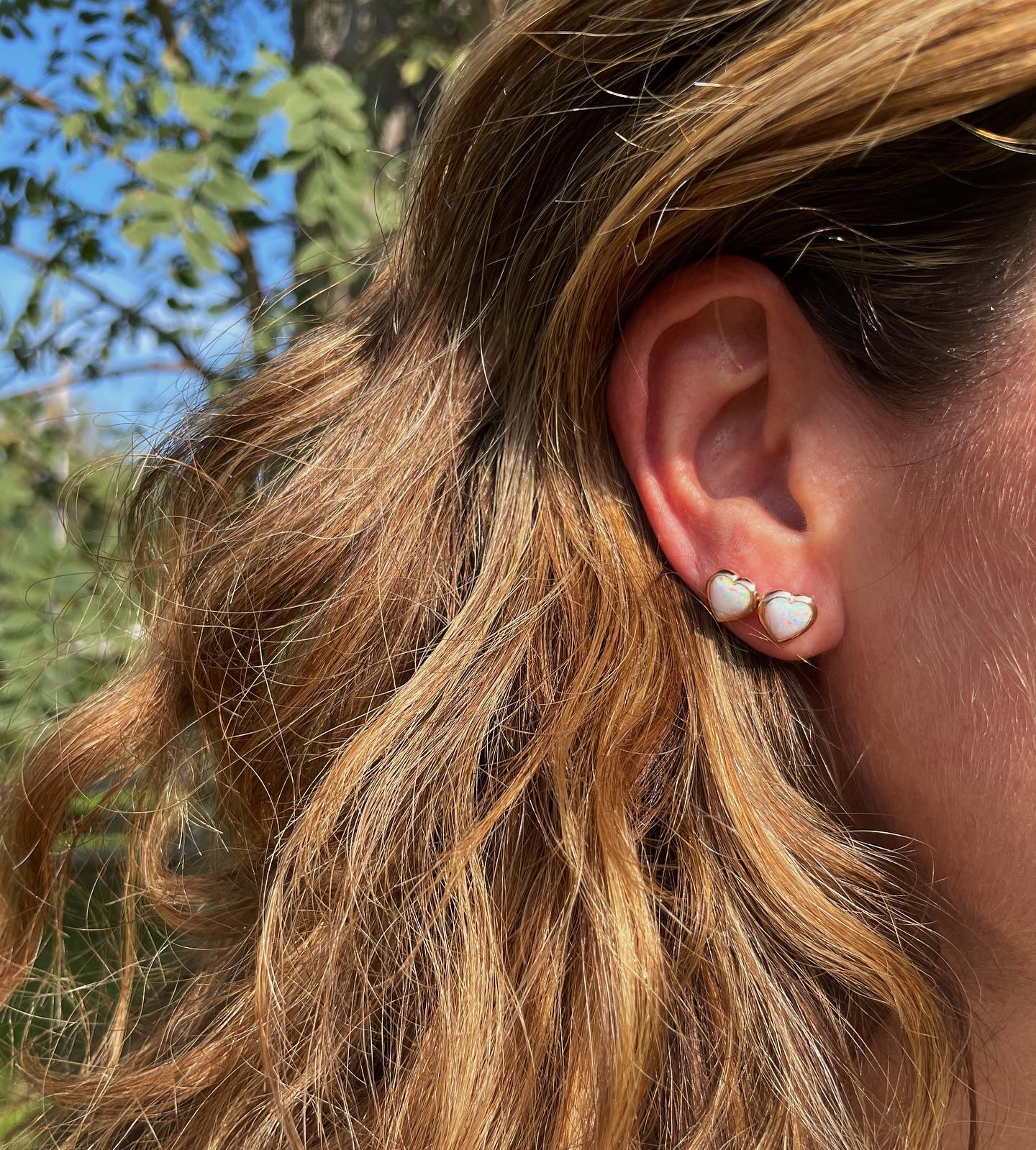 Chunky yellow gold heart shaped bezel set earrings with opal gemstones shown on ear
