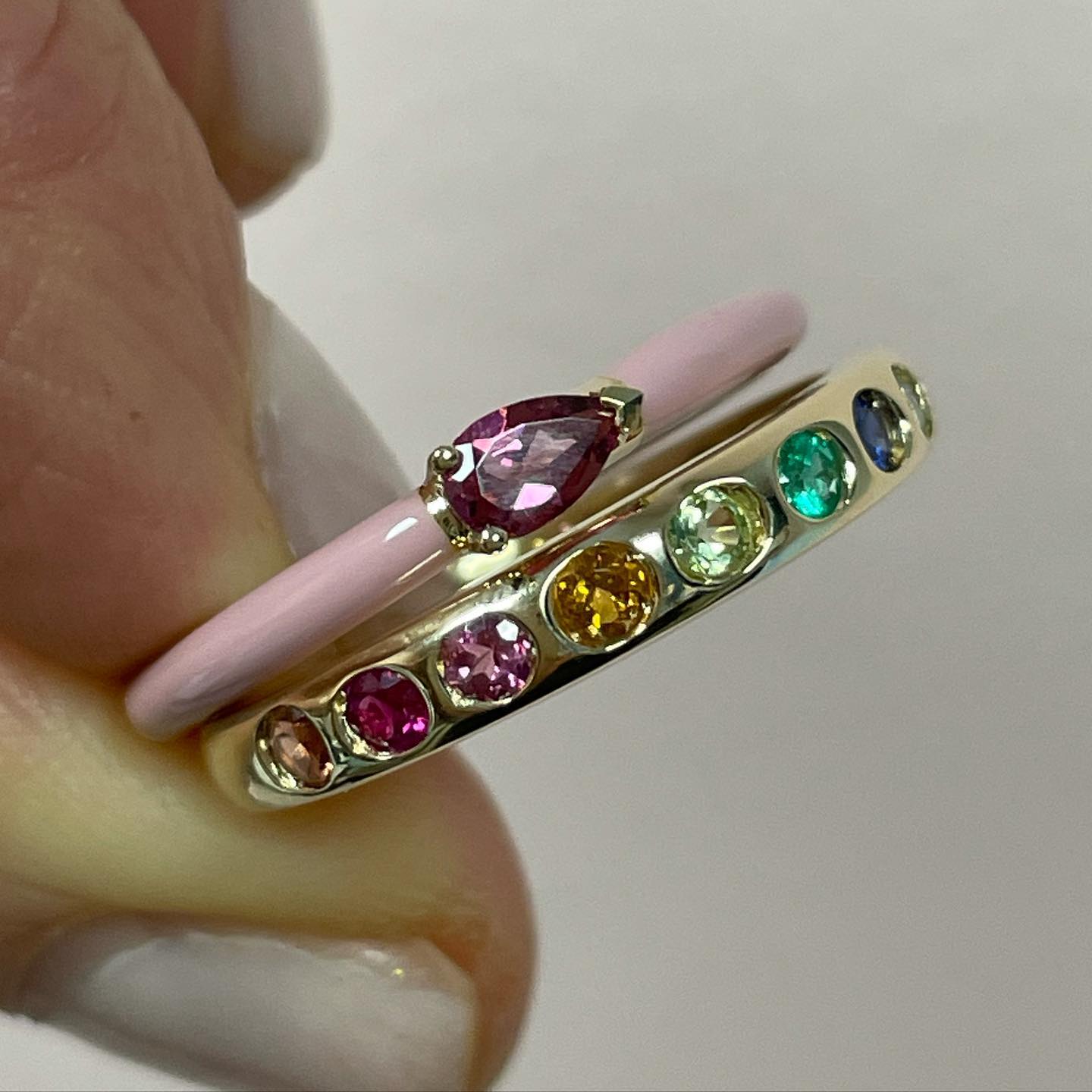 Skinny banned yellow gold ring with round multi color gemstones shown with beautiful pink enamel ring